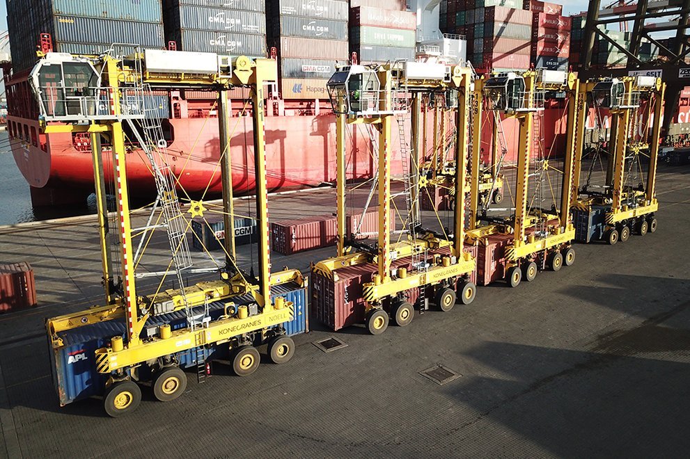Vier Konecranes Noell Straddle Carrier nebeneinander. Dahinter sind viele Container.